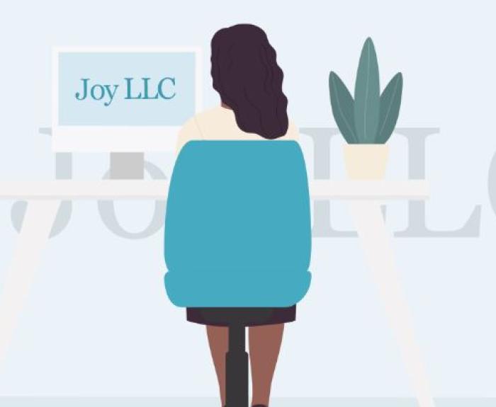 A dark haired woman named Joy sits at a white desk in a blue chair with the words "Joy LLC" on her computer monitor. There is a plant on her desk.